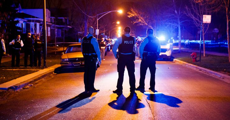 Three police officers stand in the middle of a street at night. Another group of officers stands on a lawn with crime scene tape wrapped around lamp poles in the background. 