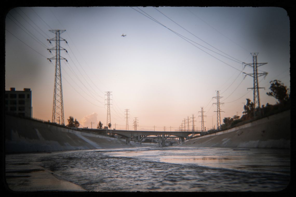 A photo shows the Los Angeles River flowing behind the Lincoln Heights Jail at sunset. The sky is visible above, in a gradation of pink, white and blue shades. A small bridge reaches across from one bank to the other. The photo has a vignette effect. 