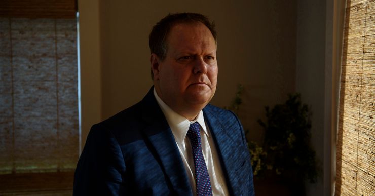 Chris Morton, a man with light-toned skin and wearing a blue suit jacket and tie, stands near a window.  