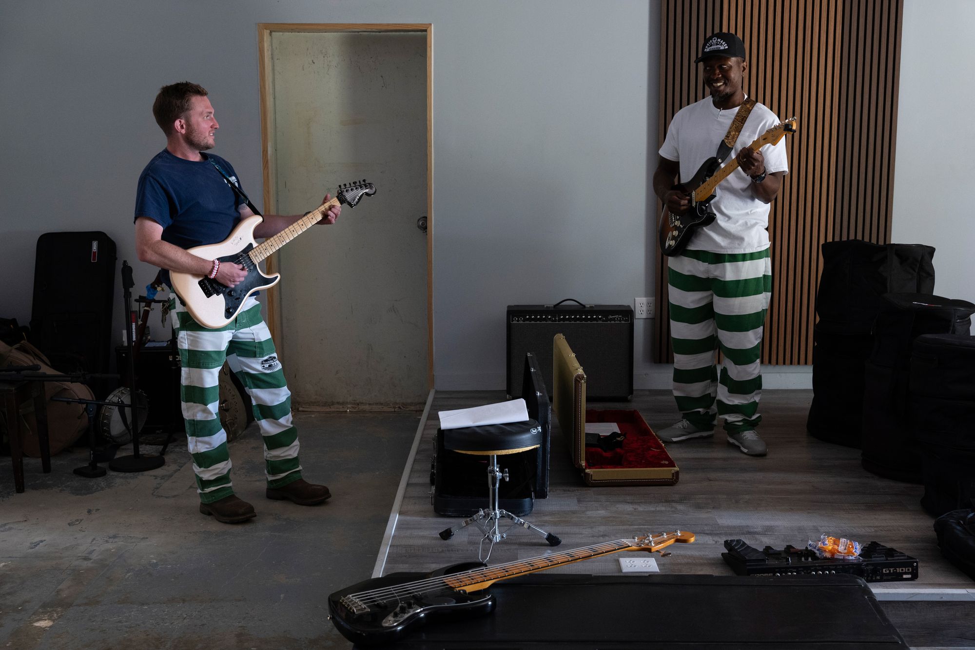 A photo shows a White man on the left, dressed in a navy blue T-shirt and green-and-white striped pants, playing a guitar; and a Black man in a white T-shirt and green-and-white striped pants, playing a guitar. They are standing in a rehearsal room with an amplifier, an instrument case, and a stool, among other musical equipment.