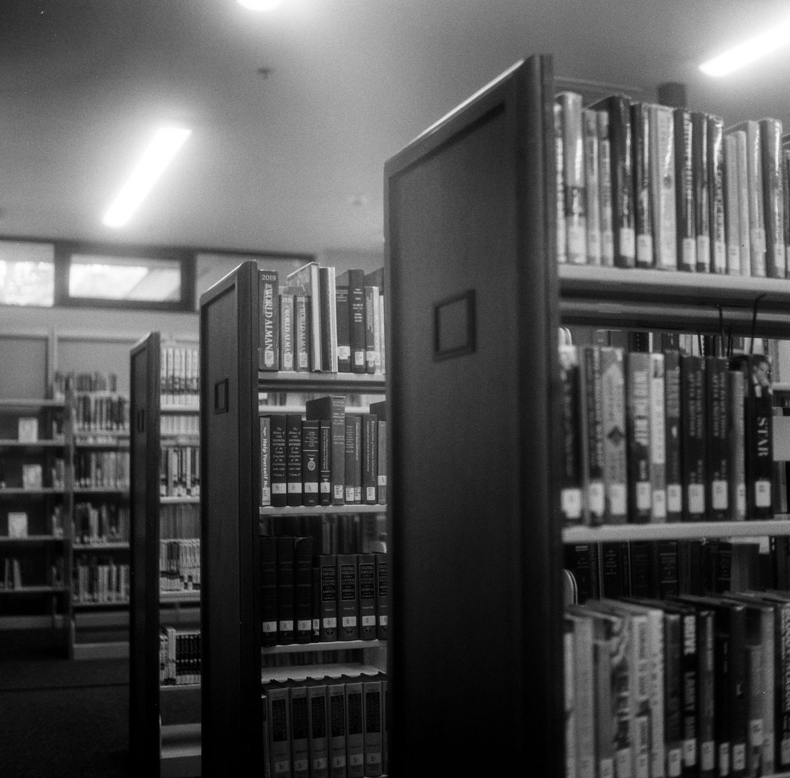 From left to right, black-and-white photos show a row of patterned chairs with wooden legs in a carpeted room, rows of bookshelves in a library, and two book drop boxes outside a library. 