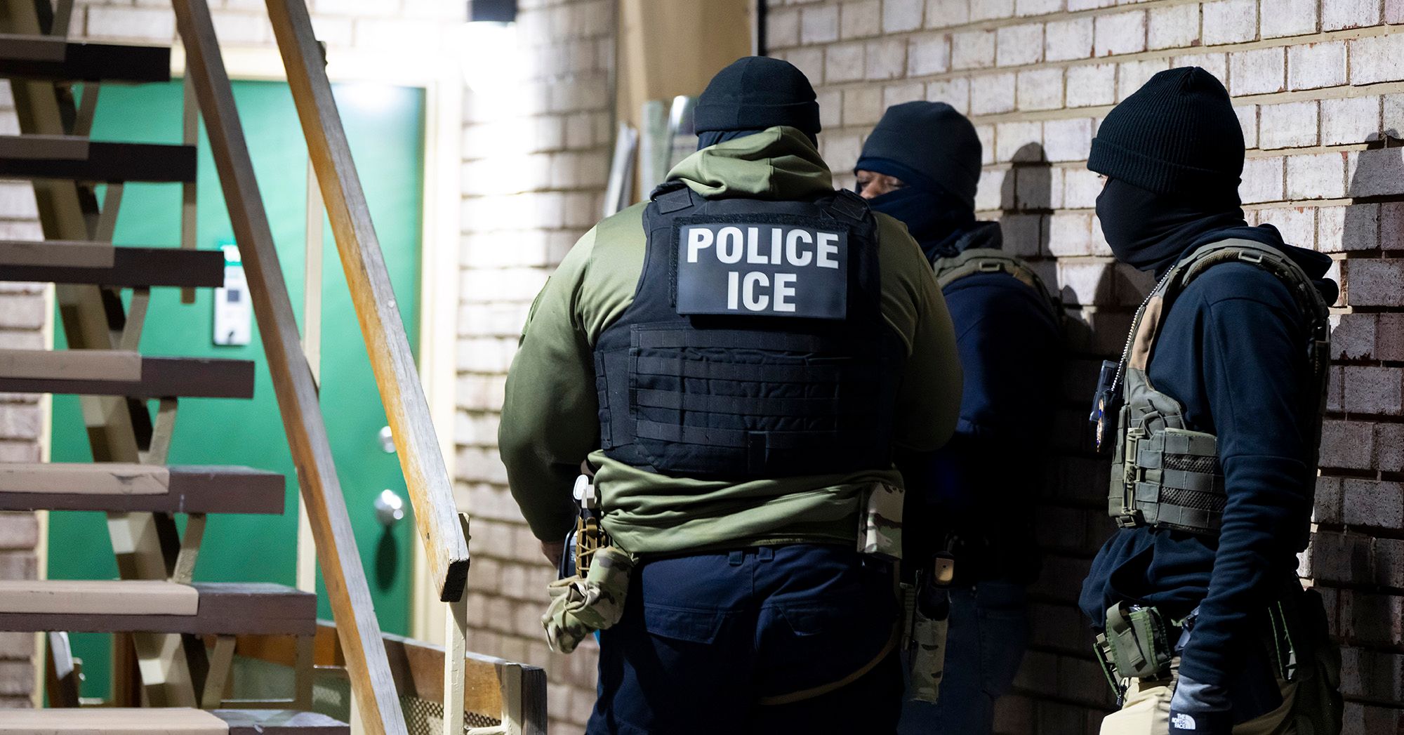 Three U.S. Immigration and Customs Enforcement officers, wearing black guard vests, green long sleeve shirts and black ski masks, stand in front of a green door.