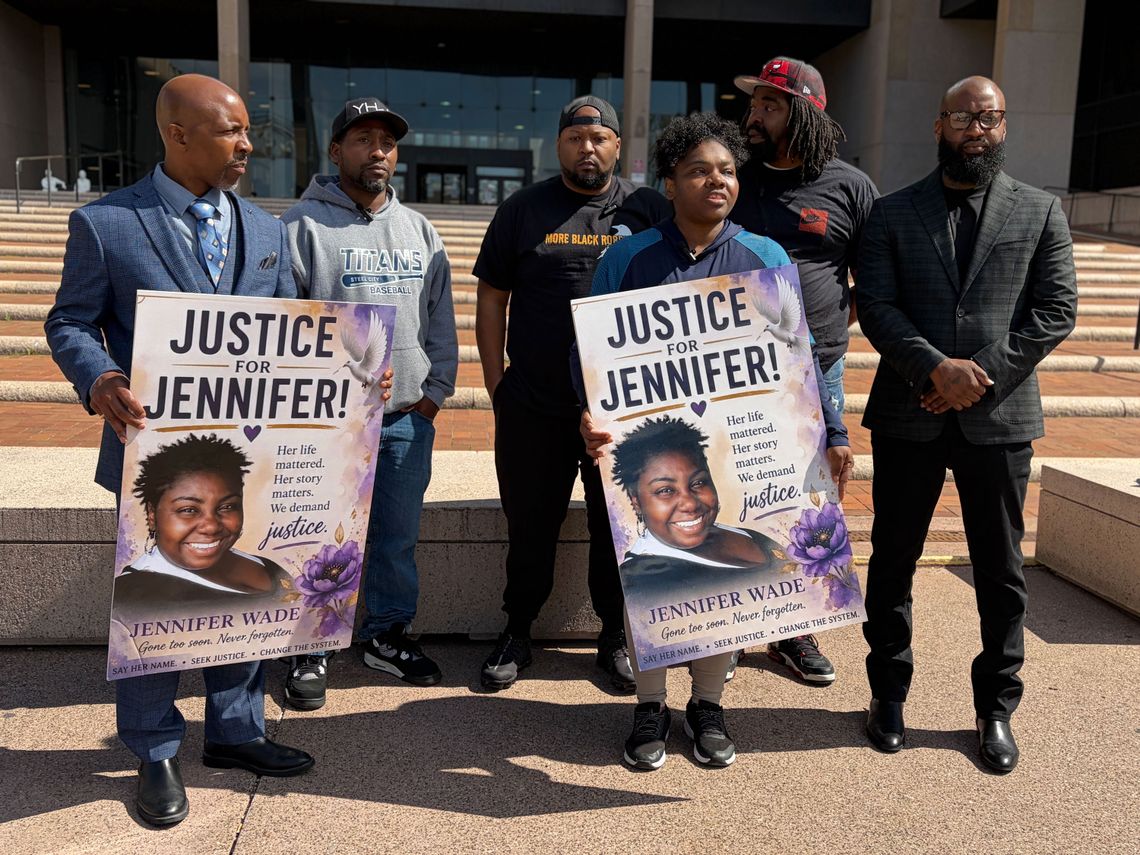A photo of a group of six middle-aged Black people in front of a building. In the front row, two people in blue, one of them a lawyer in a suit, hold signs that read, "Justice for Jennifer!" with a photo of a smiling Black woman. 
