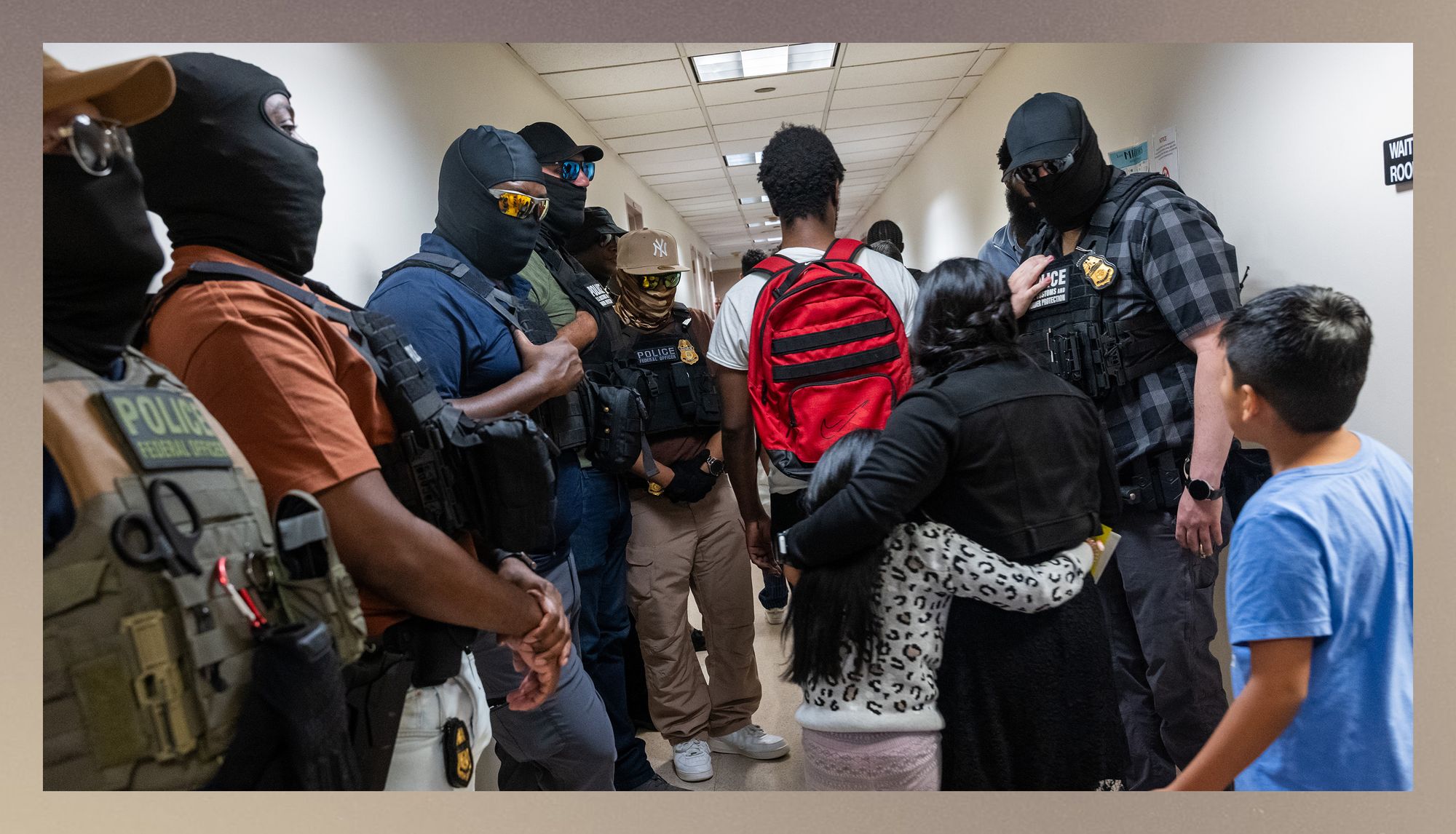Masked federal agents, including members of ICE, stand on either side of a hallway. A person wearing a white T-shirt and red backpack walks down the center of the hallway with their back to the camera. A woman and a young child, both with dark hair, walk behind the person with their arms around each other. A young boy with dark hair and a blue T-shirt follows behind them. 