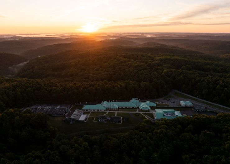 An aerial photo shows the campus of a residential treatment facility with a light green roof during sunset. The facility is surrounded by forest and hills.