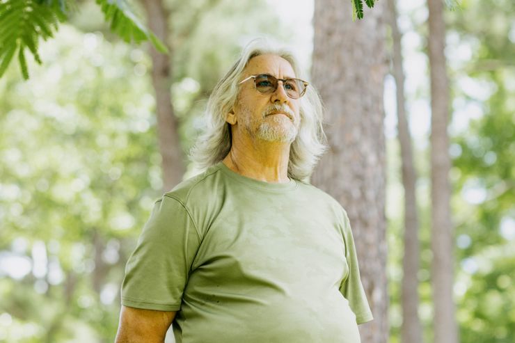 A photo of John Cook, a White man with gray shoulder-length hair, a gray beard, glasses and a green shirt. Cook is looking into the distance while standing outdoors under a tree. 