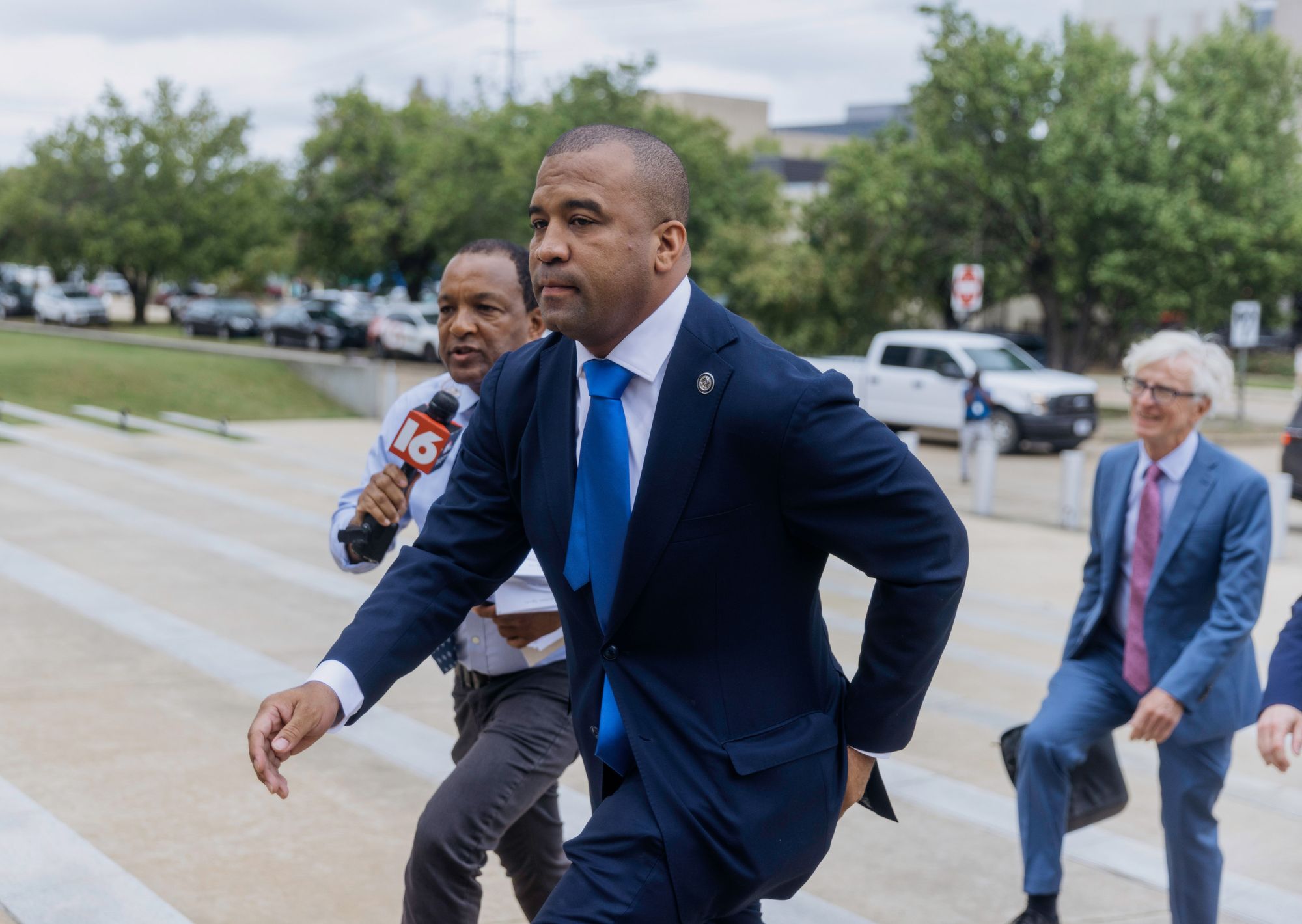 A Black man in a navy suit and blue tie walks up the steps outside of a courthouse, next to a Black man with a TV news microphone, in front of a White man wearing a blue suit and holding a leather briefcase.