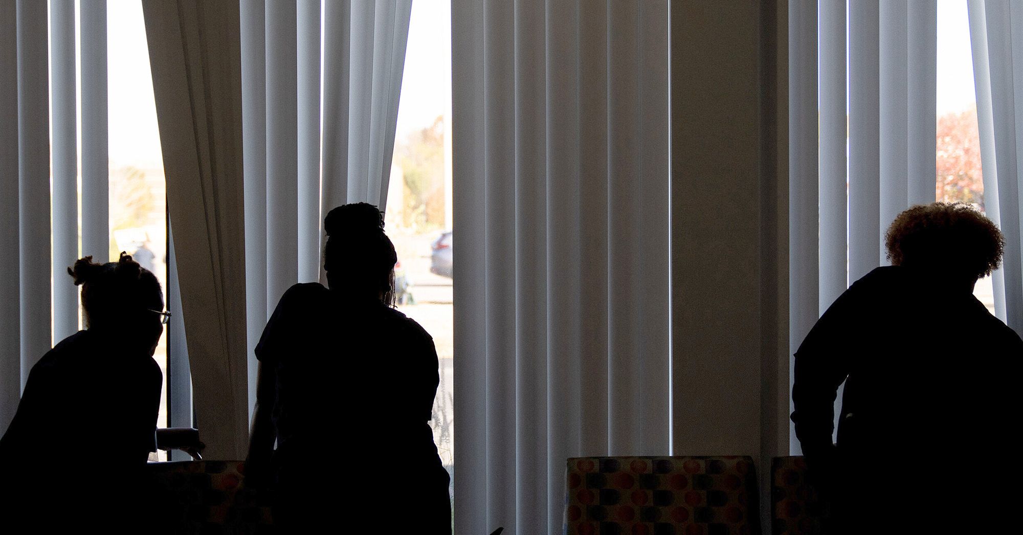 Silhouettes of staff members looking through window blinds to view a parking lot.