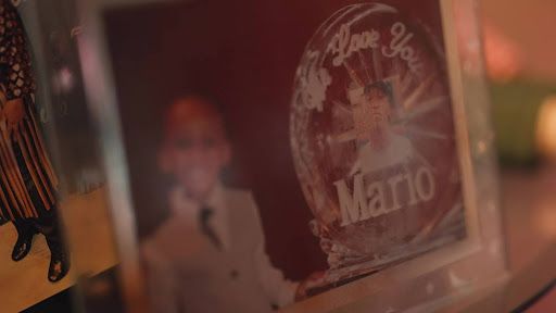 A photo of a smiling young Black boy wearing a dark tie and gray vest inside a silver picture frame alongside a photo of him as a young man with the words “We Love You Mario.”  