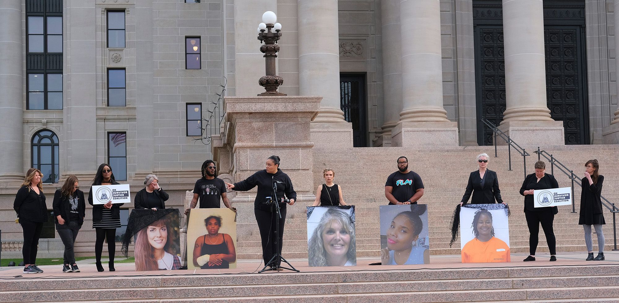 Demonstrators, dressed in black clothing, hold large posters of women and signs that read "OK Survivor Justice Coalition" while standing outside the Oklahoma State Capitol. 