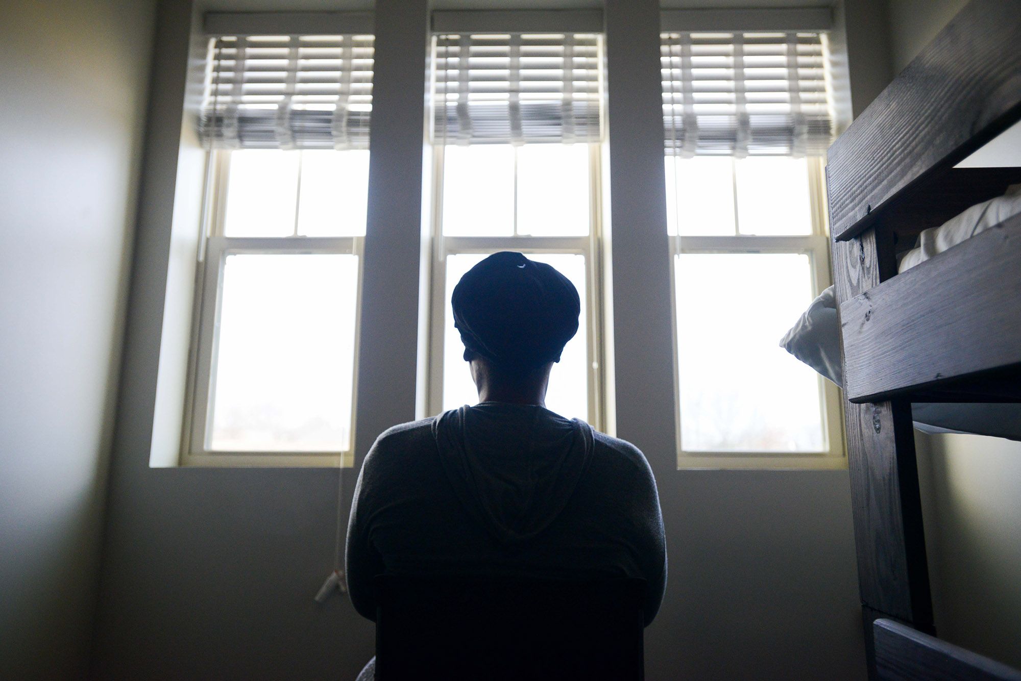 A photo shows the back of a woman as she looks out of a window. The wooden frame of a bunk bed is next to her on the right. 