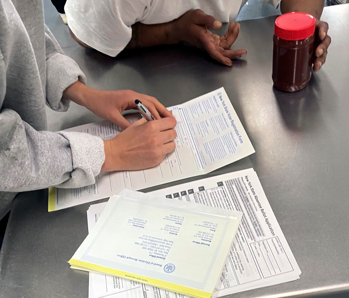 A light-skinned person in a gray sweater writes on a voter registration form next to a Black person in a prison uniform.