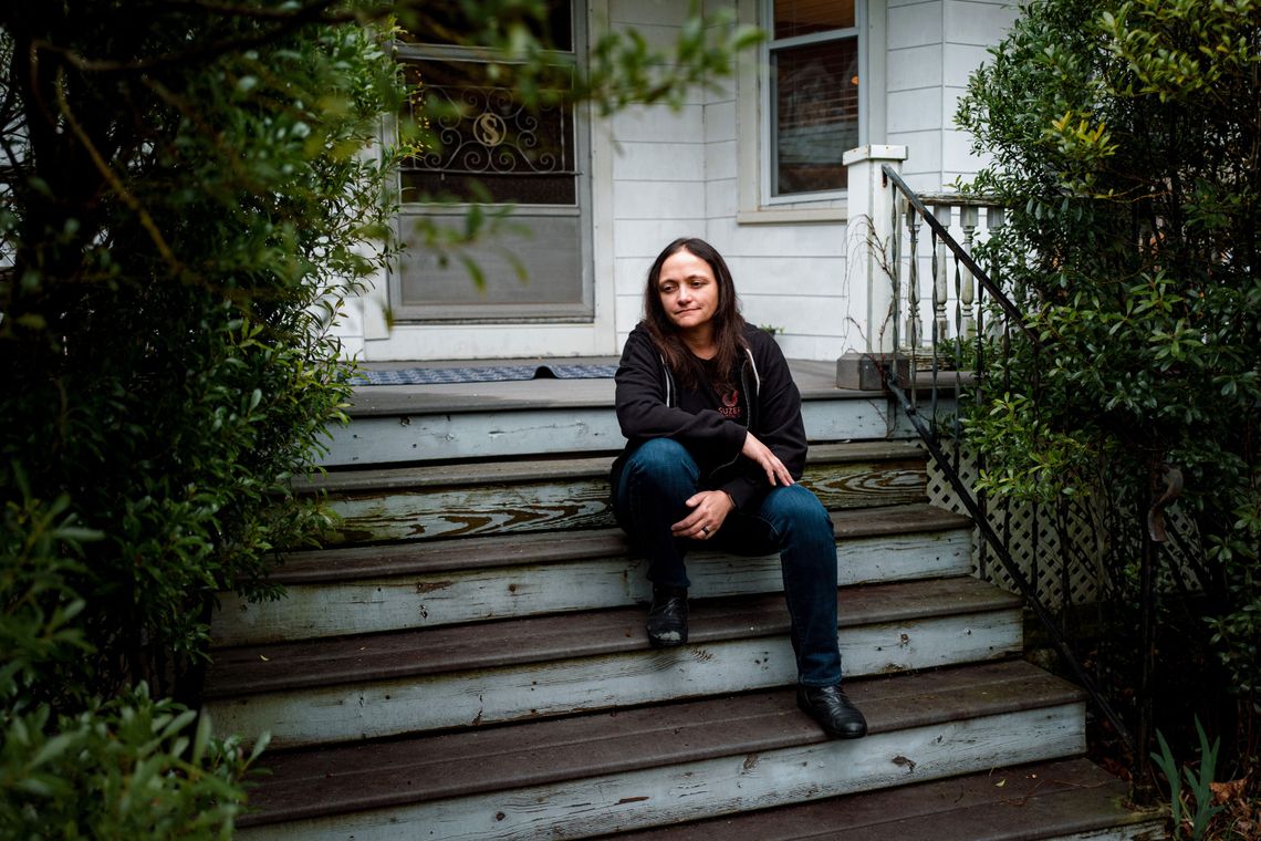 A photo of a White woman with brown hair sitting on the steps leading up to the porch of a white house, while looking to the side.