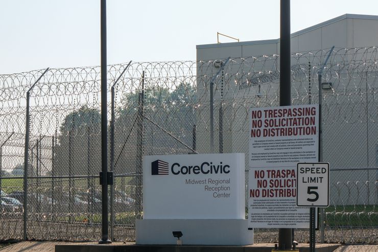 A white sign with the words “CoreCivic Midwest Regional Reception Center” is in front of barbed wire and a white building.