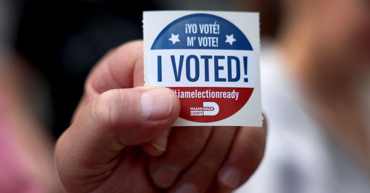 The hand of a person with light-toned skin holds a red, white and blue sticker that reads “Yo voté!” “M’ vote!” “I voted!” and “iamelectionready.”