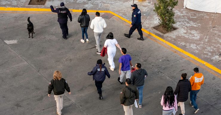 A photograph with an aerial view shows a group of eight people walking on a road as they follow several immigration officers.