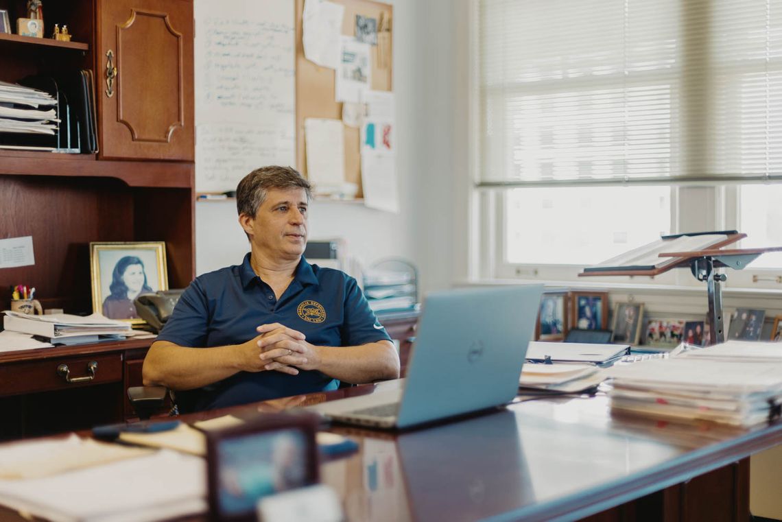 A White man, wearing a blue polo shirt, sitting in front of his office desk.