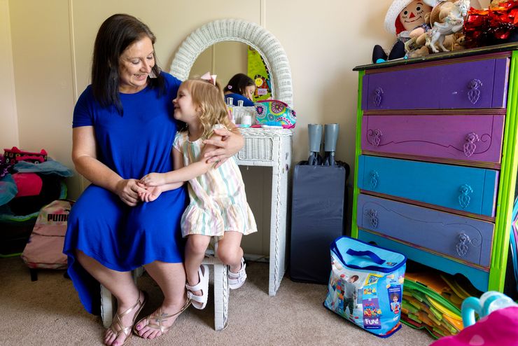 A photo of a White woman with shoulder-length brown hair and a blue dress posing with her daughter, who is wearing a striped pastel dress and a bow in her blond hair. The woman and her daughter are sitting in a child’s room. 