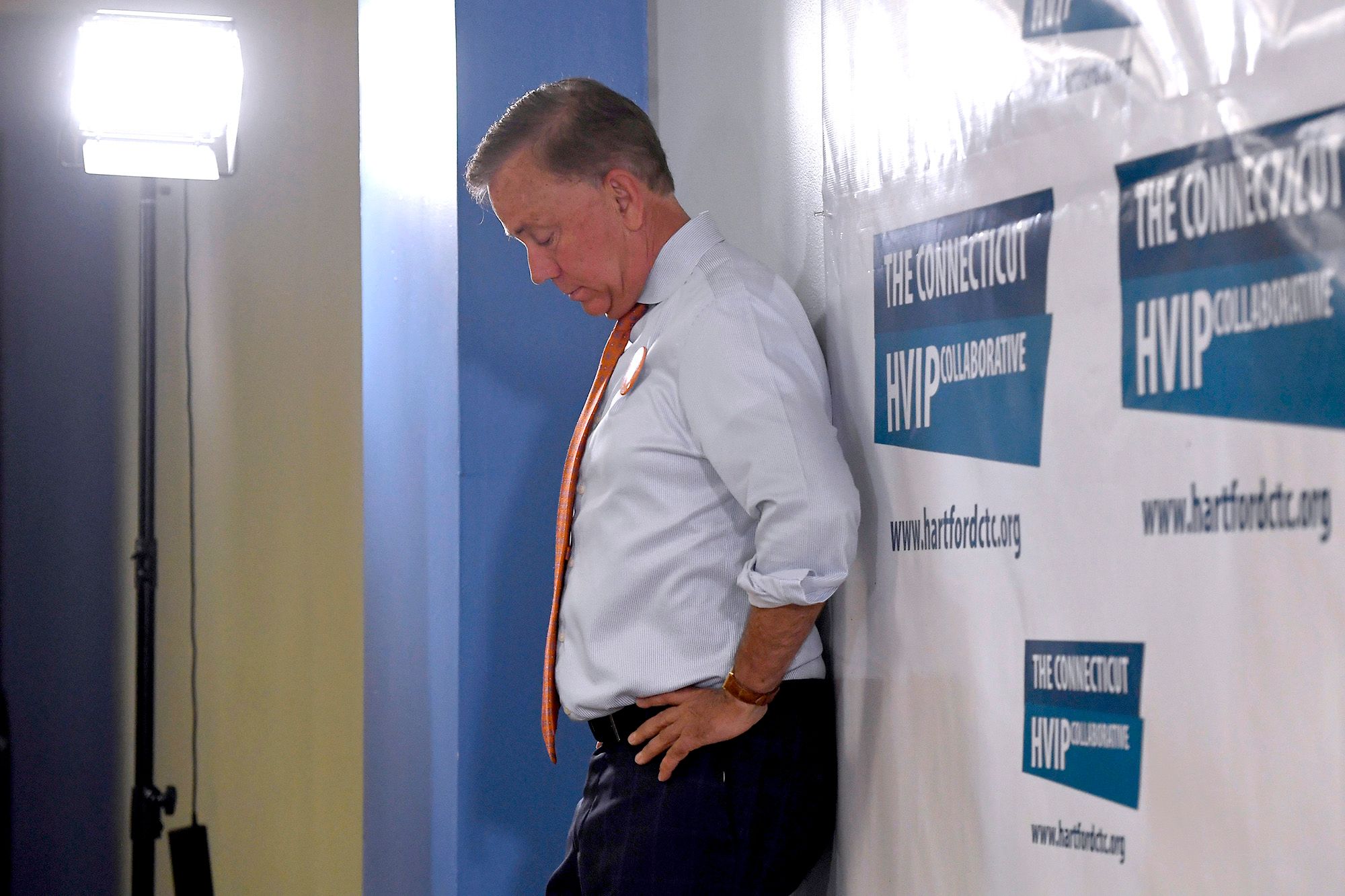 An older White man is looking down with his hands on his hips, while leaning against a backdrop that says "The Connecticut HVIP Collaborative." He is dressed in a white button-up shirt with rolled-up sleeves, an orange tie and black pants. 