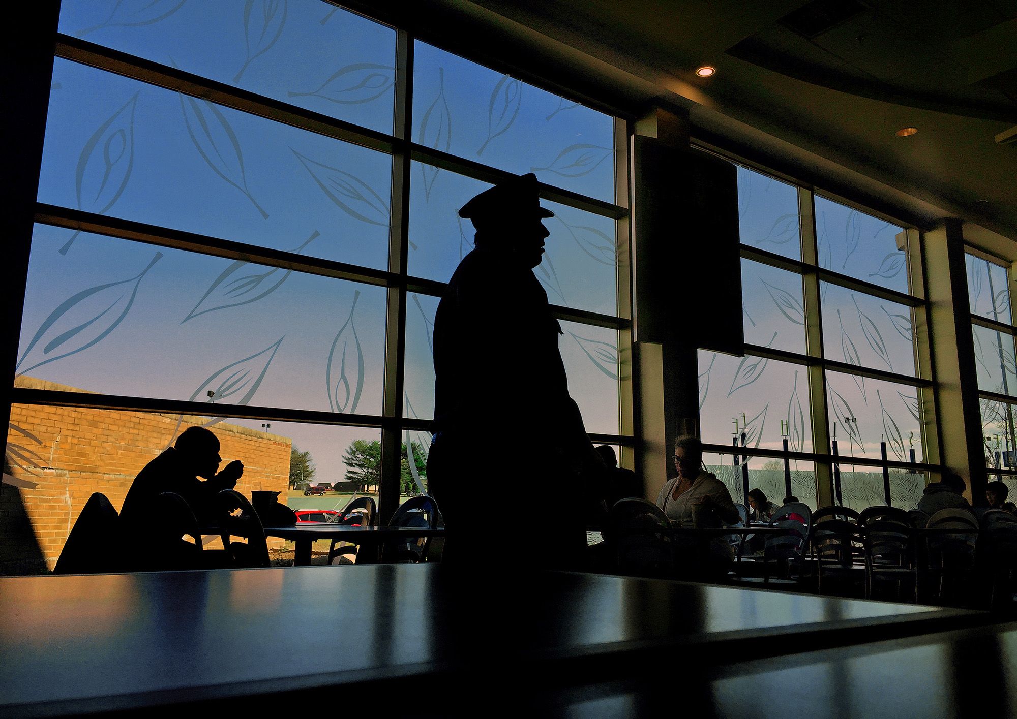 A silhouette of a security guard standing in the middle of a mall's food court. In the background, people are eating while sitting at different tables. 