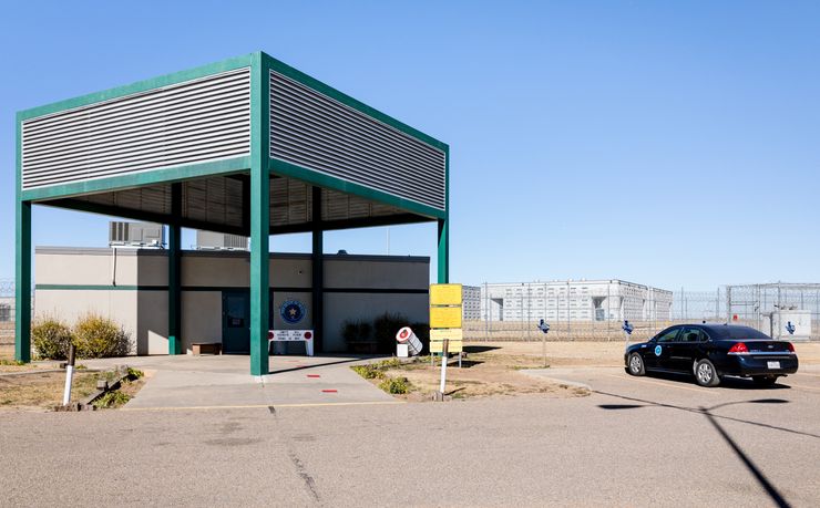 The entrance to a Texas prison facility, a gray rectangular building with a detached teal shaded structure in front of it. A black car is parked to the right. There are wired fences and buildings in the background. 