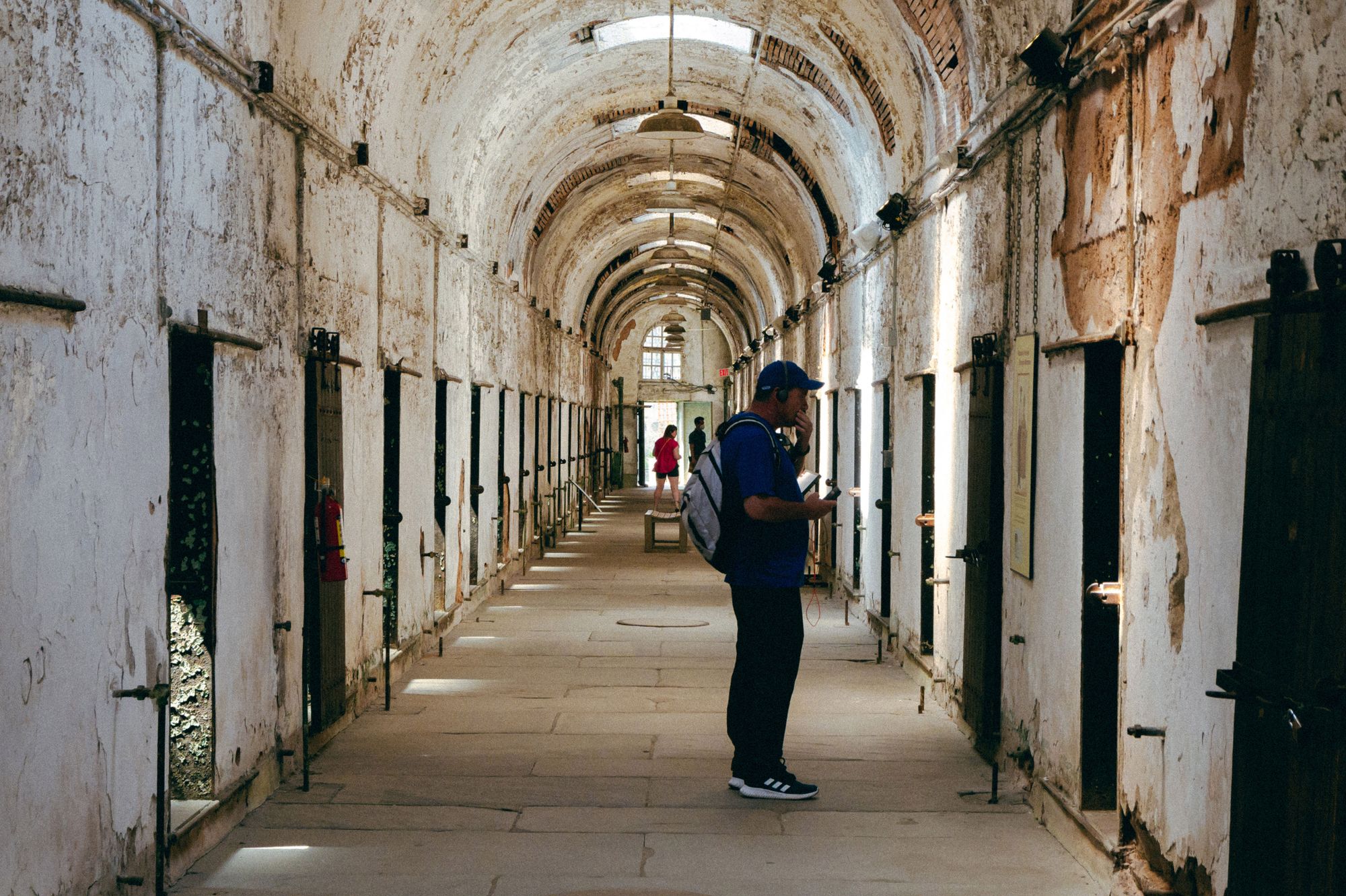 A man in a blue shirt and black pants looks at the door of a prison cell, which is one of many on either side of a long hallway with a rounded ceiling. 