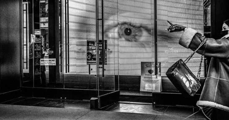 A black and white photograph shows a large electronic billboard with a closeup of an eye at the front of a store in New York.  