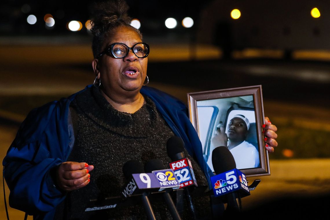 A Black woman with glasses, holding a portrait of her son, speaks in front of several news mics. 