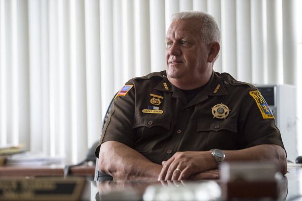 Frederick County Sheriff Chuck Jenkins, a White man, sits in a brown sheriff's uniform at his desk in his office. 