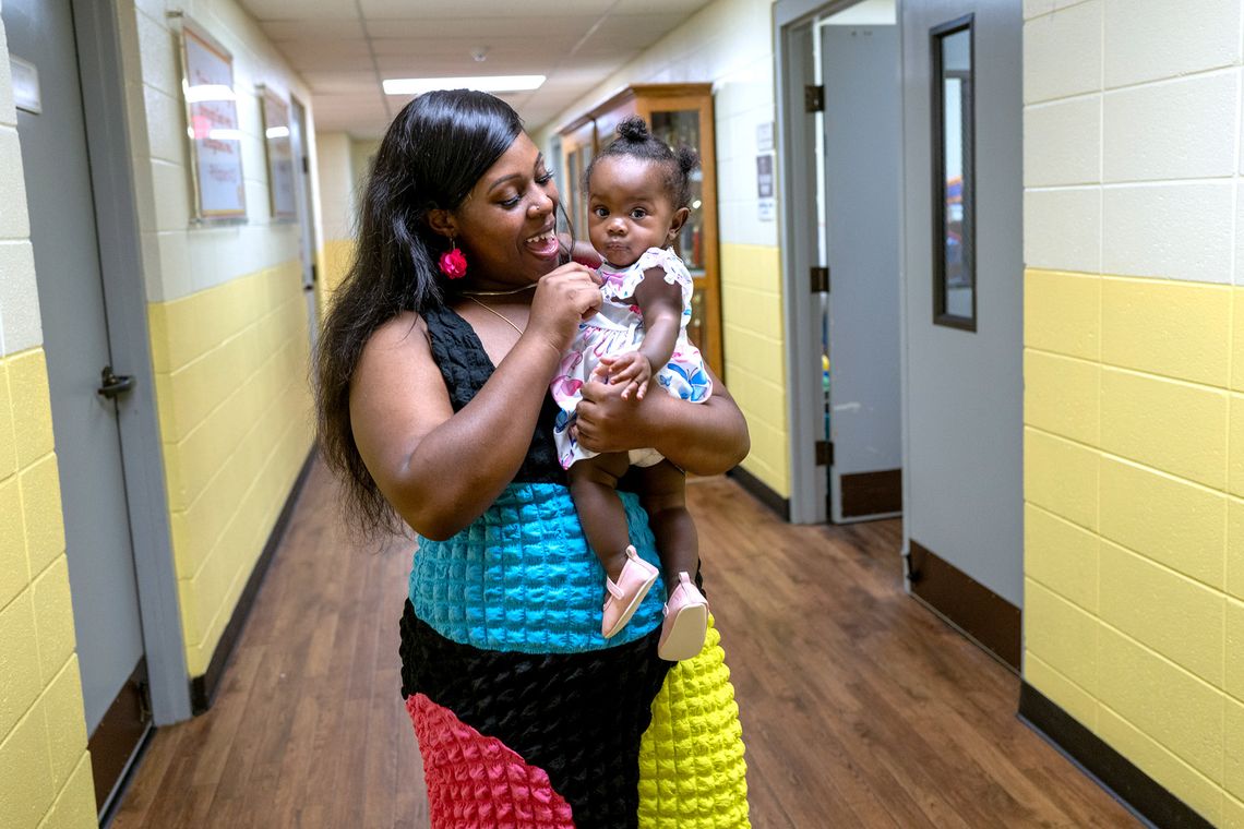 Melissa Robinson, a Black woman wearing a teal blue, magenta, black and yellow dress, smiles as she holds her daughter. 