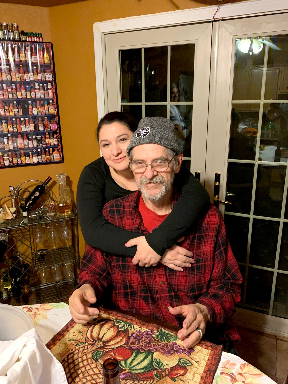 A light-skinned woman stands behind an elderly man with her arms around him, while he sits at a dining room table. 