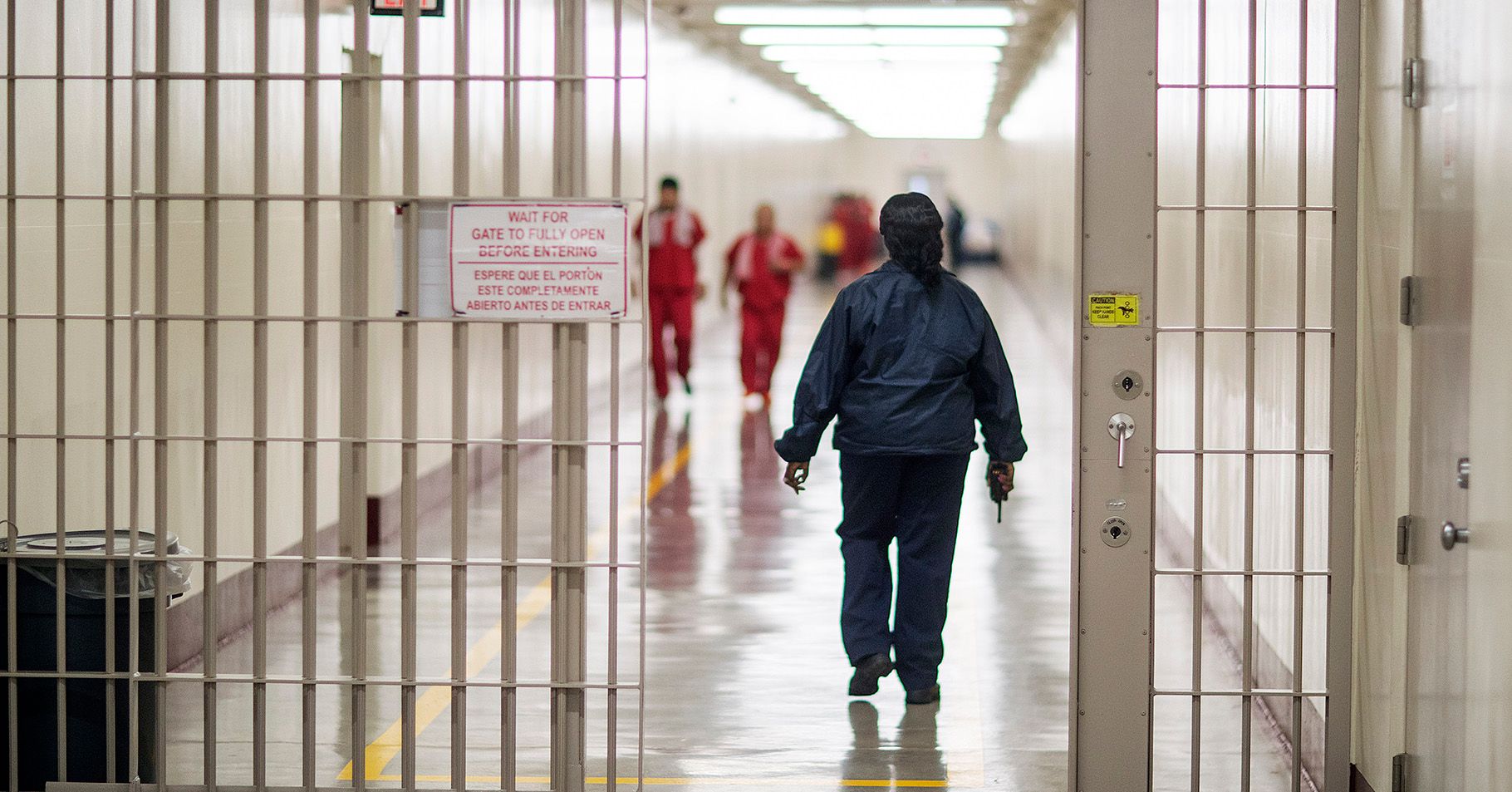 An officer in a navy uniform holds a walkie talkie while walking through a hall. There is an open gate in the foreground. Two people in orange uniforms with towels are walking in the background.