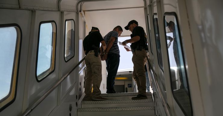 A man looks down with his arms behind his back, as he is being handcuffed by a police officer at the top of a jet bridge walkway. An officer to the right reads from a piece of paper. 