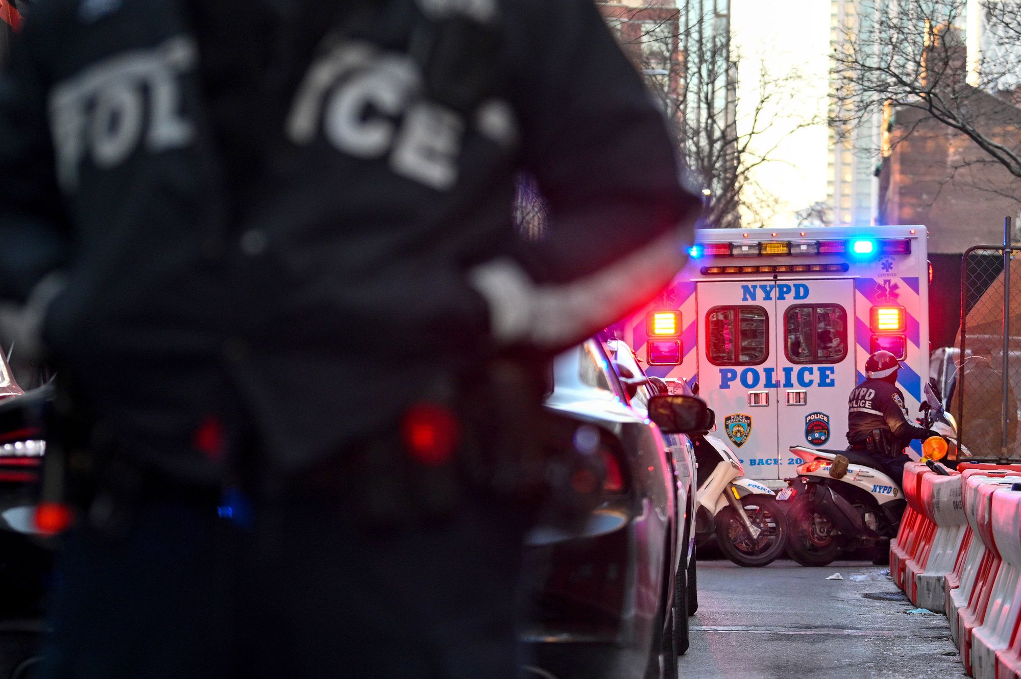 A New York Police Department ambulance is parked on the right, with an officer on a motorcycle behind it. A officer's chest is out of focus in the foreground on the left. 
