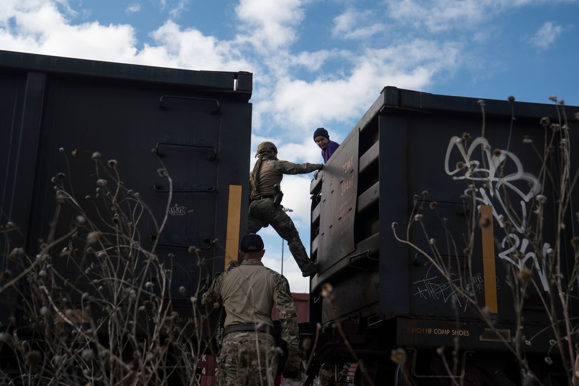 A woman in a purple shirt looks over the top of a train car at two Texas Department of Public Safety special operations agents. One agent has climbed partway up the train car to speak with her. 
