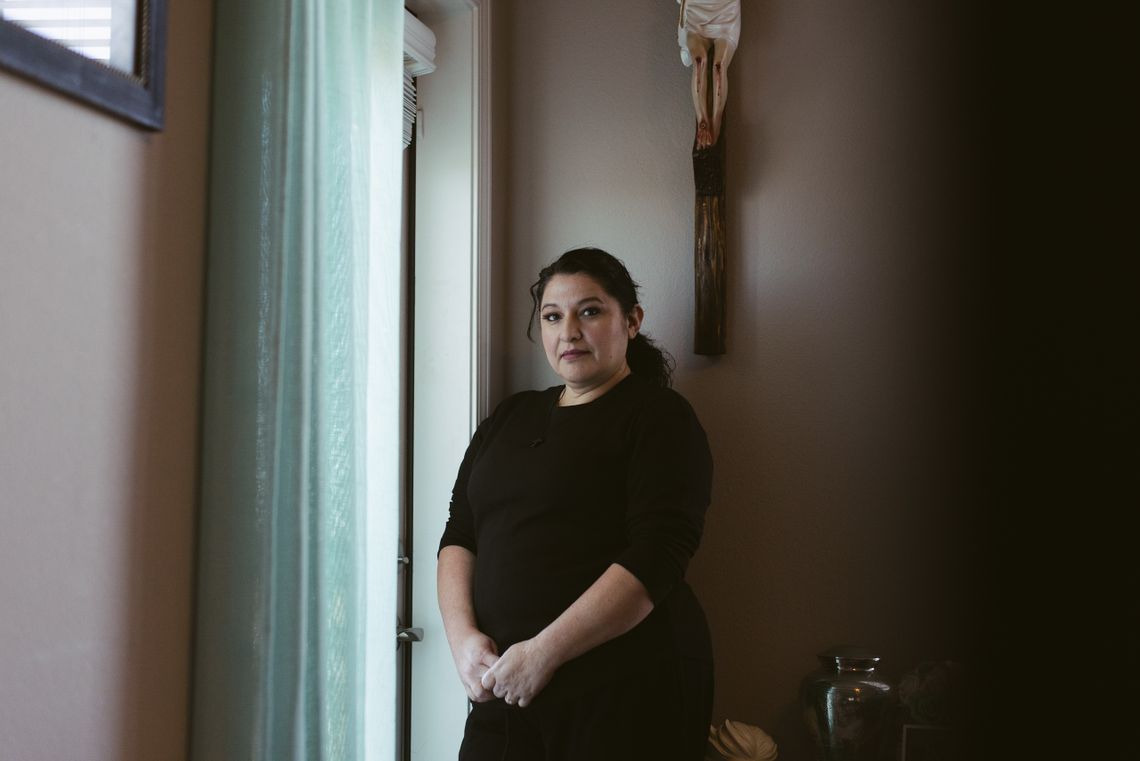 A light-skinned woman in a black dress stands by an entrance in her apartment, staring at the camera. In the background, a cross with a sculpture of Jesus hangs from the wall and an urn is on a table. 