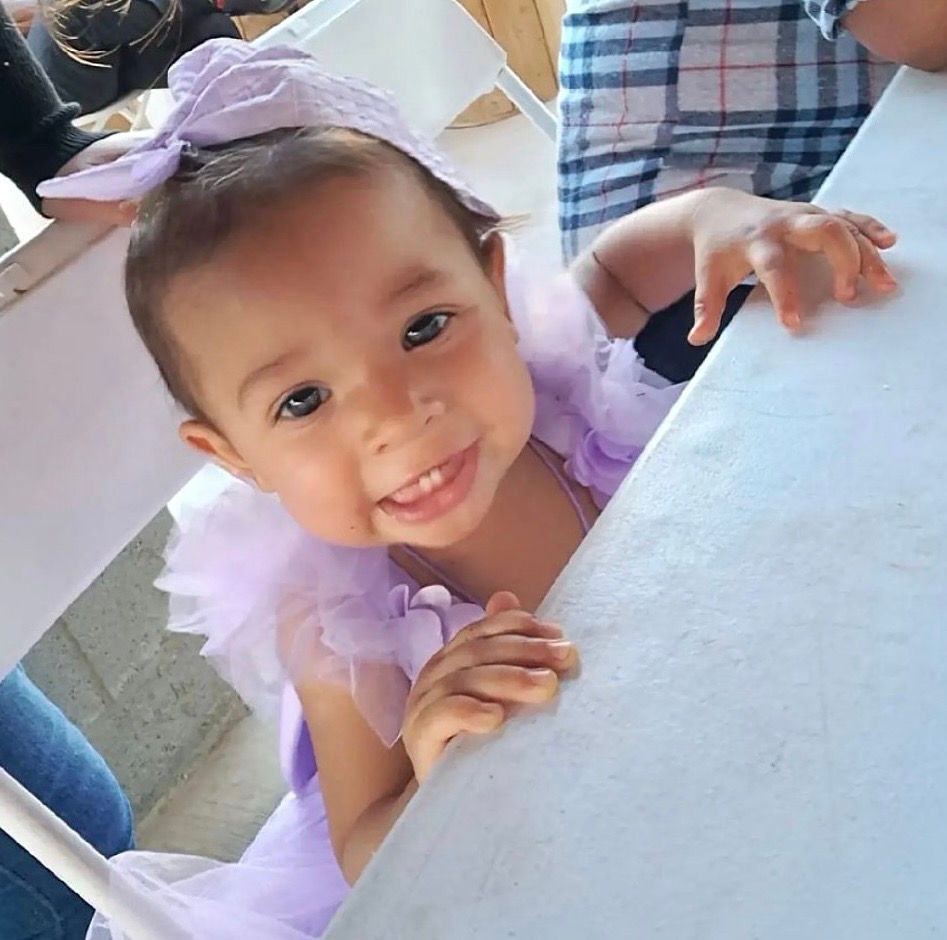 A photo shows a baby with medium-toned skin and dark brown hair tied with a purple bow, smiling as she leans on a table. 