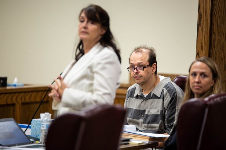 In a courtroom, a White man sits at a table peering over thick-rimmed black glasses. He is dressed in a gray-striped shirt. To his left is a woman standing in a white suit jacket and to his right is another woman, seated at the table next to him.