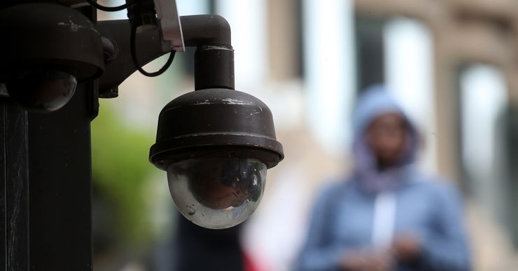 A photo shows a video surveillance camera in focus. A person in a light blue sweater is walking down a street in the background.