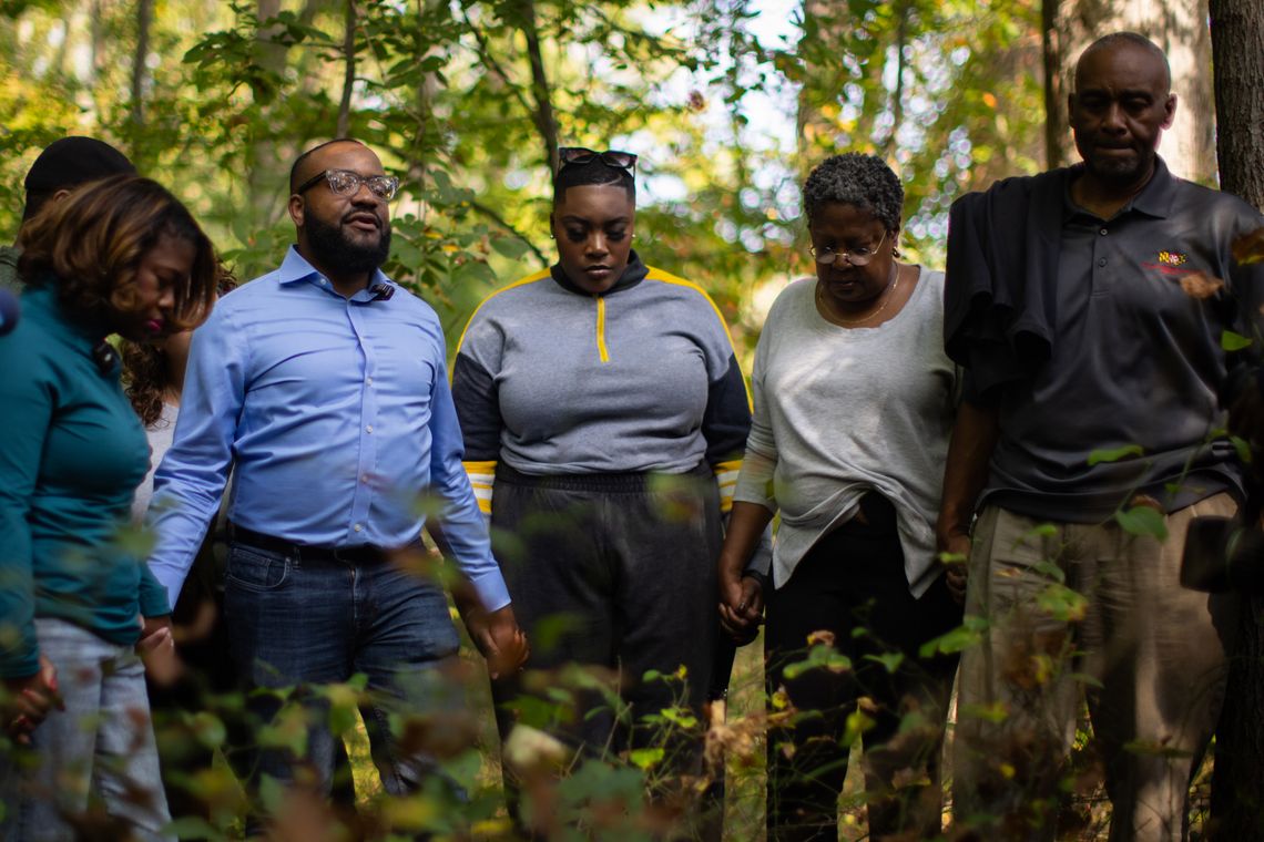 A photo shows a group of Black men and women holding hands, closing their eyes, and bowing their heads in prayer in a forest.