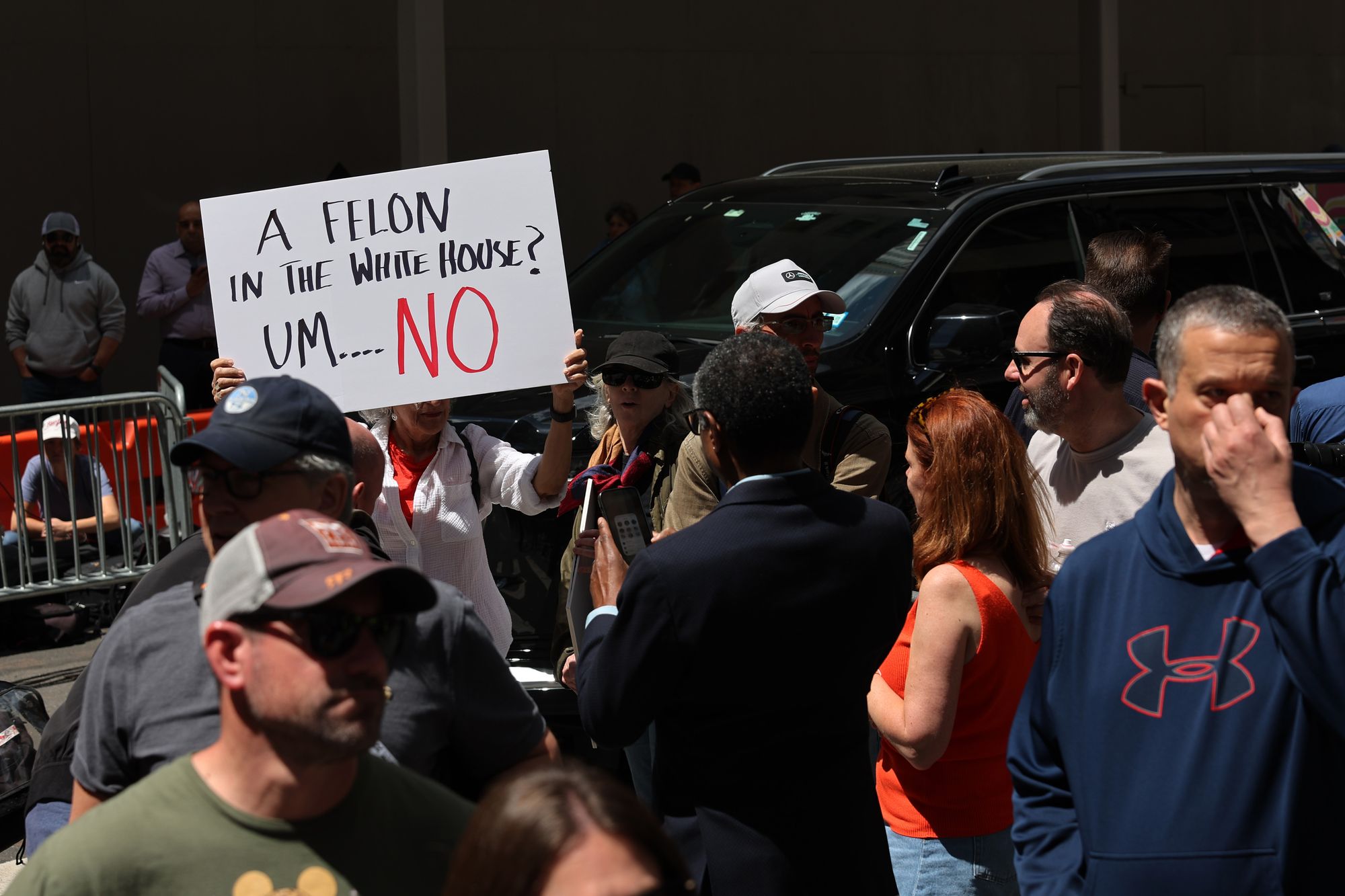 A group of people stand outside, with a vehicle nearby.  A person holds a sign that says: "A felon in the White House? Um...NO". 