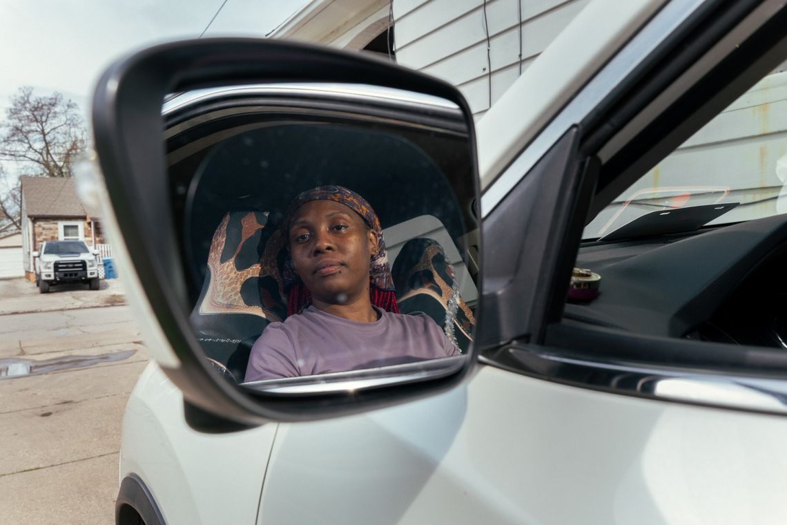 A photo shows Carmisha Bell, a Black woman wearing a purple T-shirt. She is sitting in the driver’s seat of a white car, and her head and shoulders are reflected in the driver’s side mirror. 