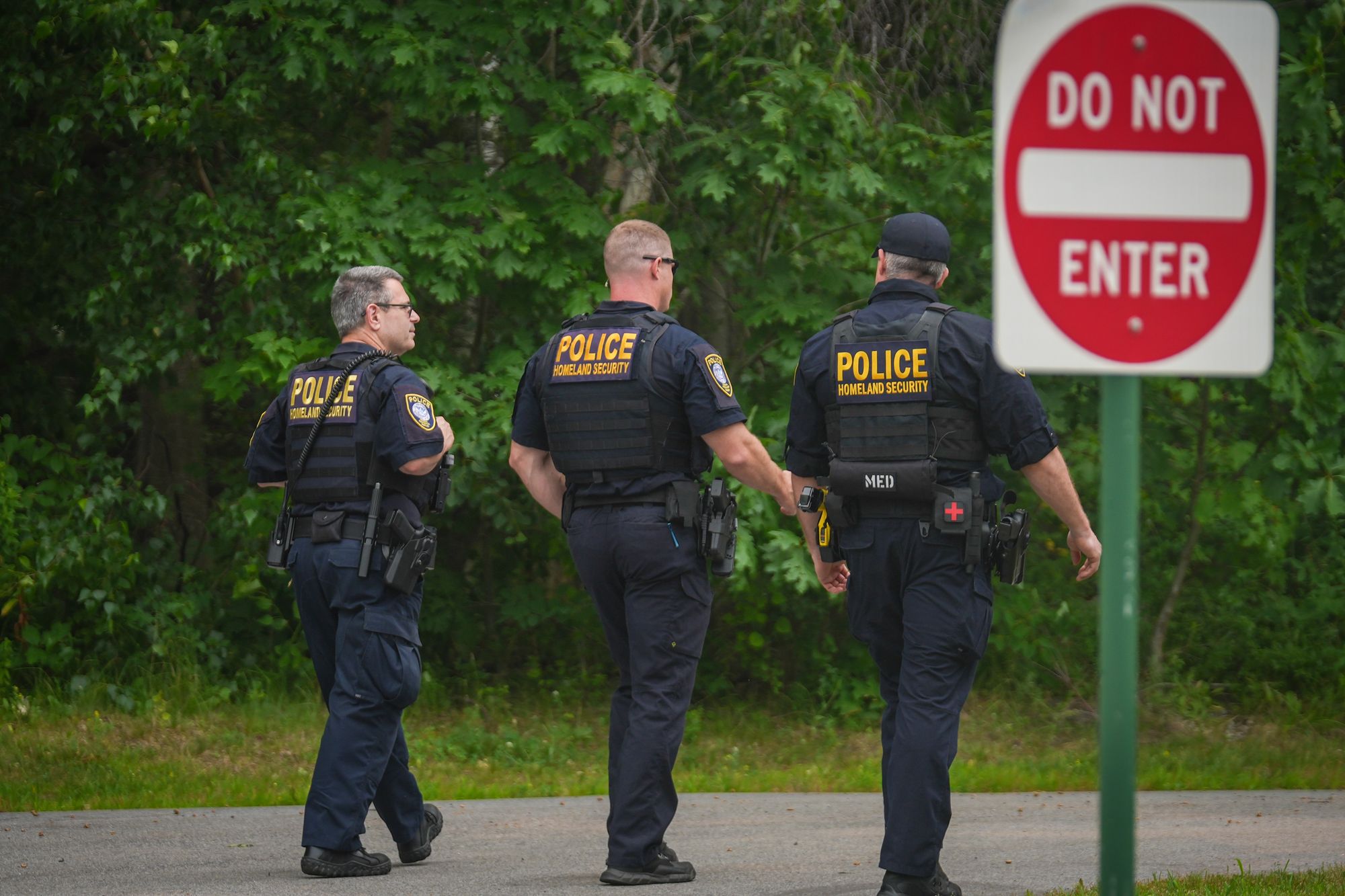 A photo shows three male Department of Homeland Security officers, dressed in dark uniforms, as they walk away from the camera.  A red and white “Do not enter” sign is visible on the right side of the photo. 