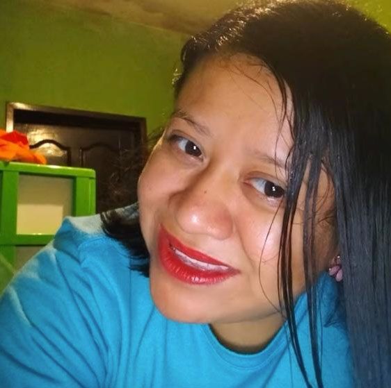 Una mujer de piel morena clara, cabello negro liso y camisa turquesa se inclina hacia la cámara para tomarse una selfie. Al fondo, una pared verde y una puerta marrón oscuro.
