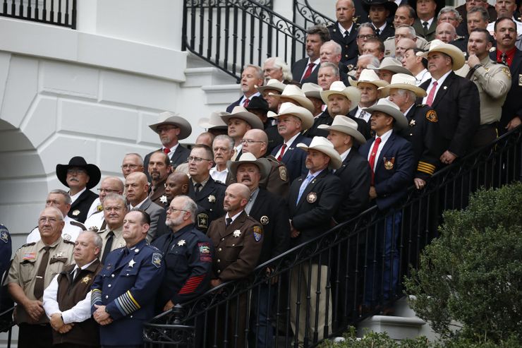 Dozens of sheriffs in uniform, some wearing cowboy hats, some wearing suits, stand on the steps of an angled staircase outside of The White House.  