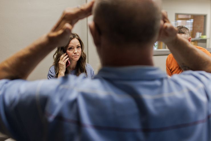 A photo of a White woman with brown hair holding a phone while talking to a White man wearing a blue shirt. The man is out of focus and seen from behind.