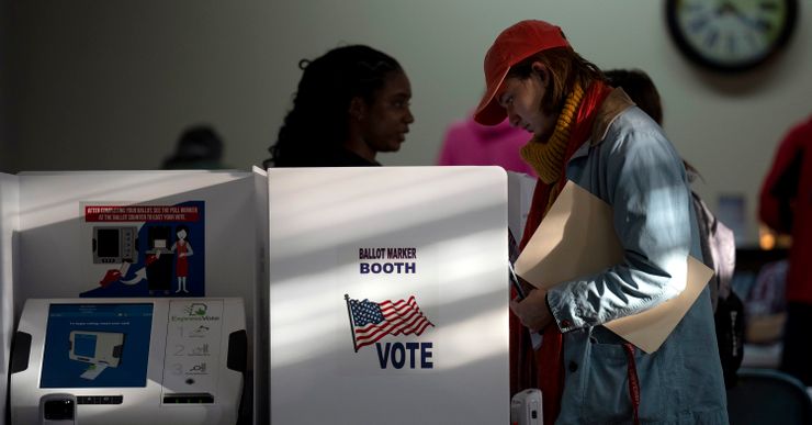 A light-skinned person, wearing a red hat and blue jacket, casts their ballot at a voting booth. An electronic voting machine can be seen on the left, in a different voting booth. 