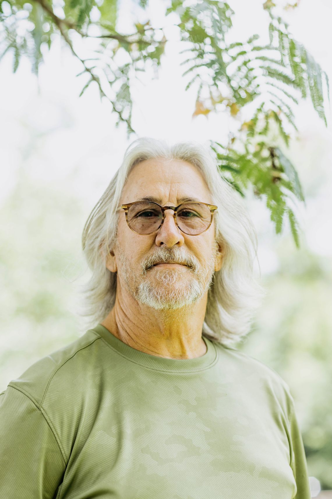 A photo of John Cook, a White man with gray shoulder-length hair, a gray beard, glasses and a green shirt. Cook is looking into the camera while standing outdoors under a tree. 