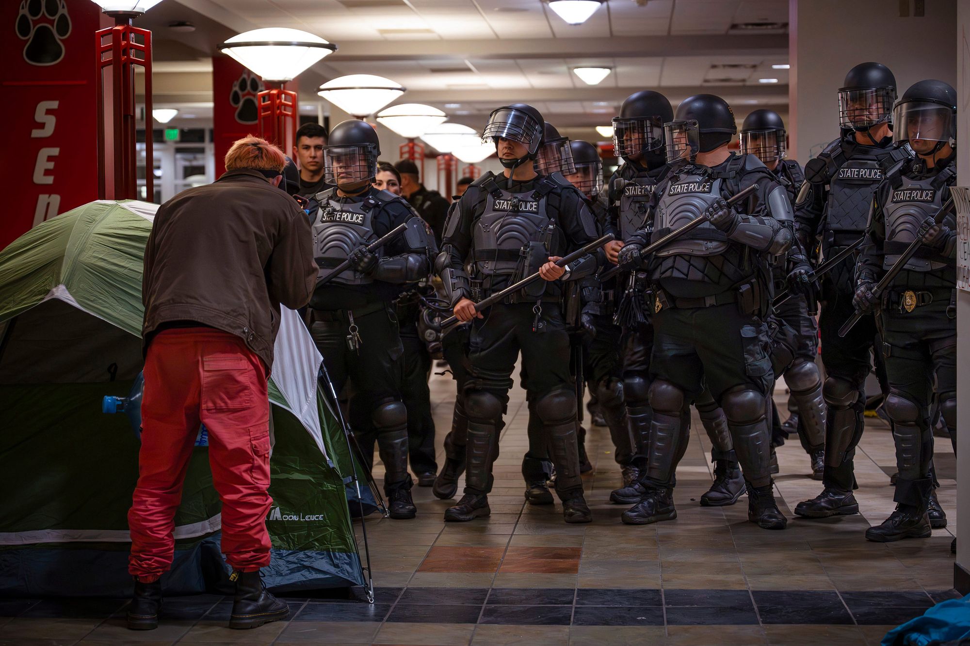 A photograph shows a group of more than 10 police officers wearing riot gear and holding batons watch a person standing by a tent.