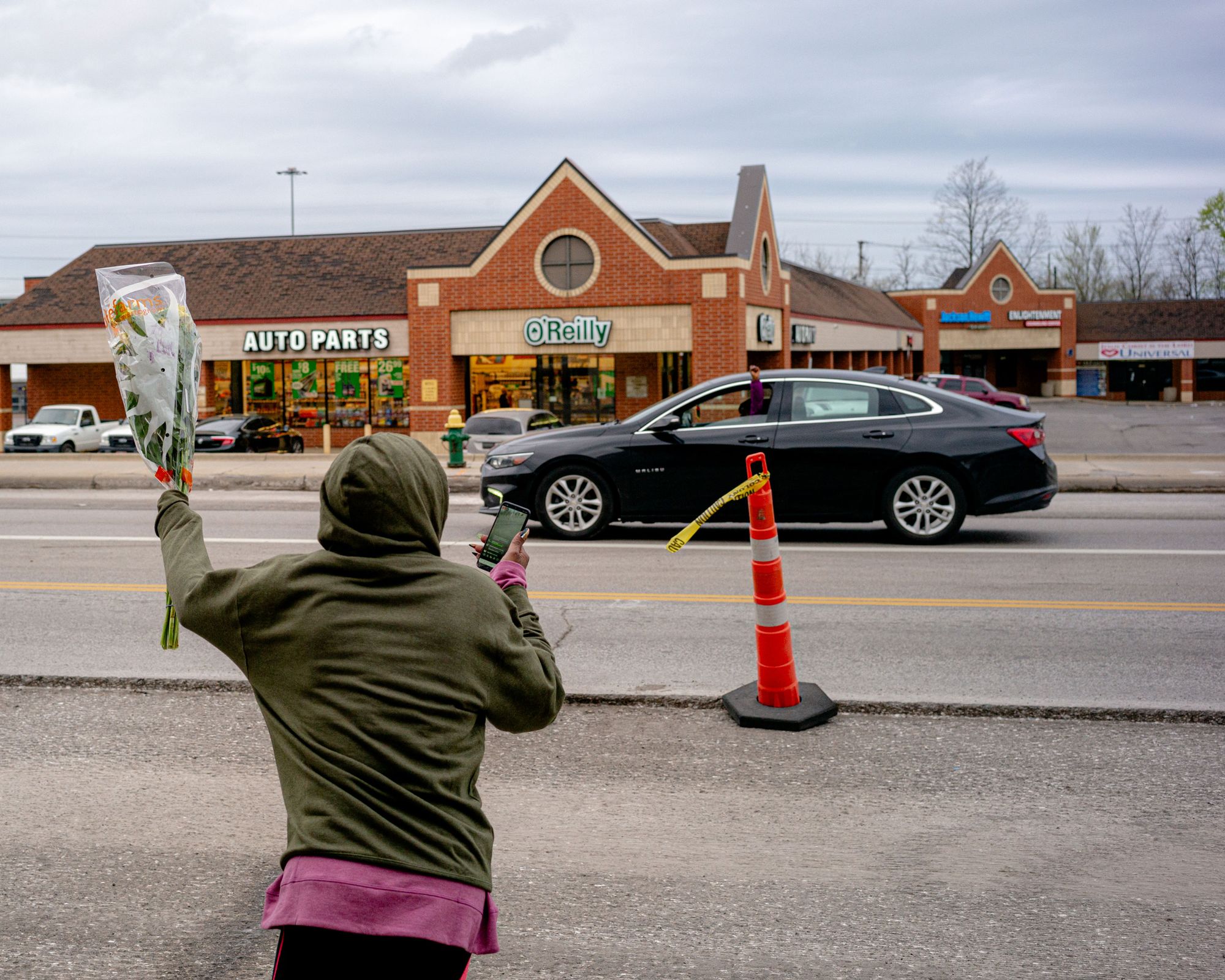 A photo of a woman in a green hoodie holding up flowers toward a passing car. 